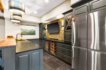 An amenity-stocked, U-shaped bar with microwave, sink, storage, refrigeration, live-edge bartop and television by Janel Swanson of Wildwood Kitchens & Baths